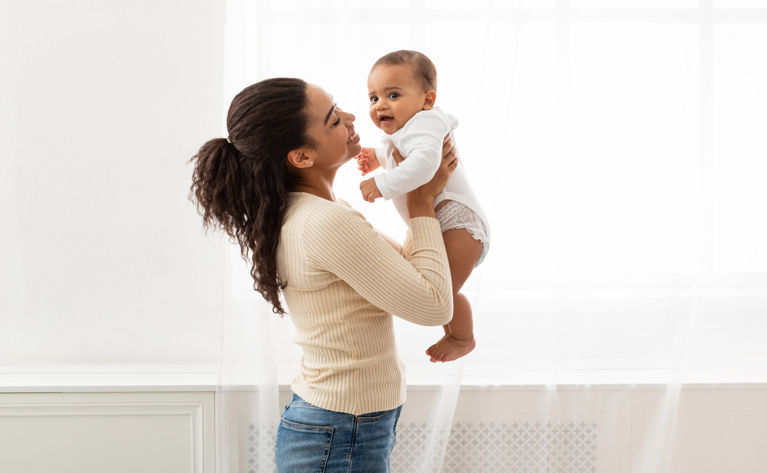 Mom's Love. Young Black Mother Holding Baby Toddler Playing And Cuddling With Little Infant Son Standing Near Window Indoor. Motherhood Happiness And Child Care, Maternity Leave Lifestyle. Side View