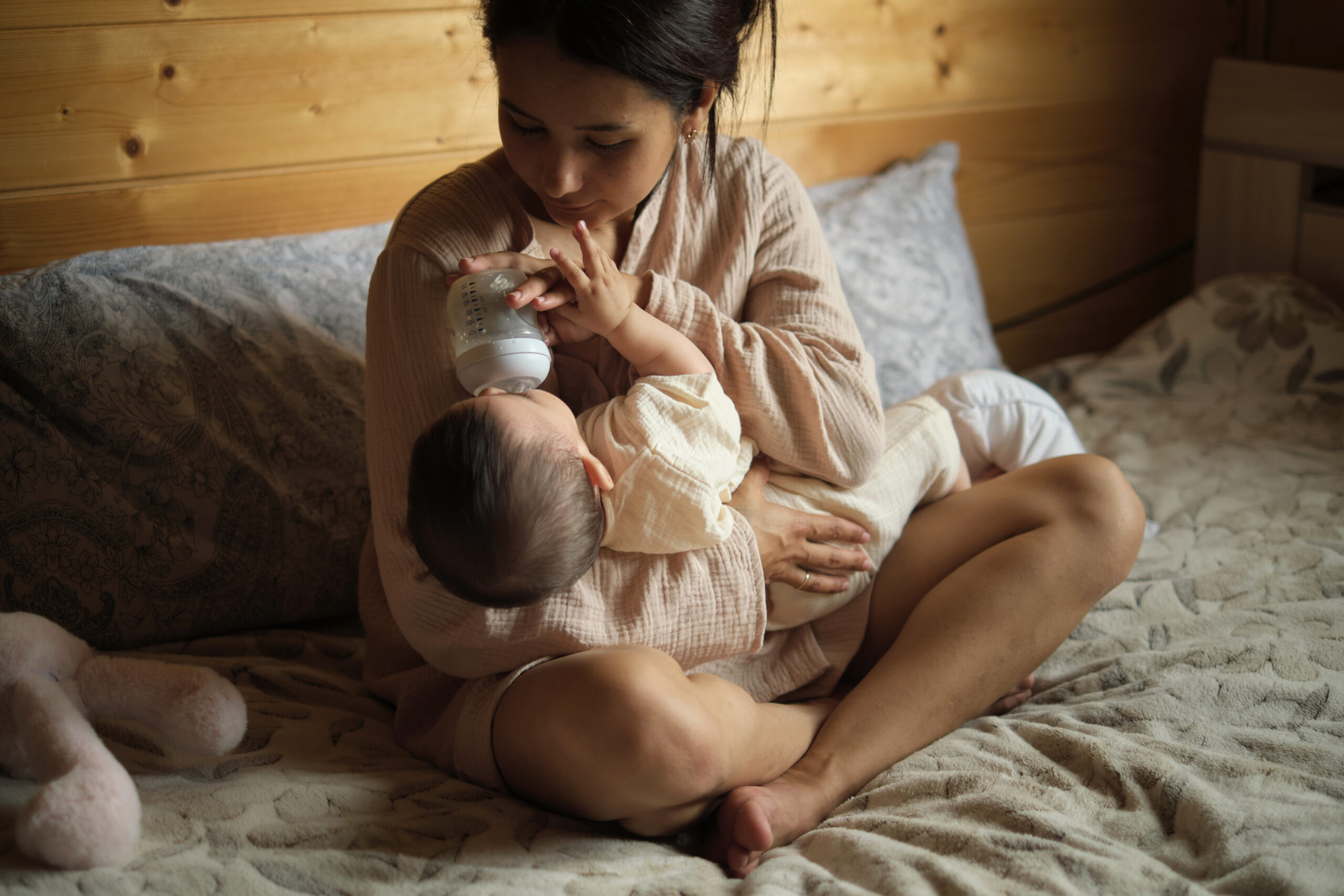 Sitting on the bed, a young mother feeds her baby with a specialized formula, demonstrating the diverse choices parents make today, sometimes opting for or relying on alternative feeding methods